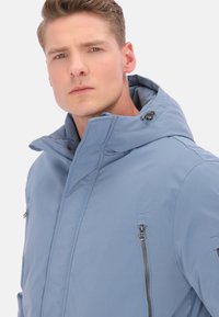 Világoskék kapucnis dzseki sima textúrával, magas gallérral, cipzáras mellzsebbel és állítható fém díszítésekkel.