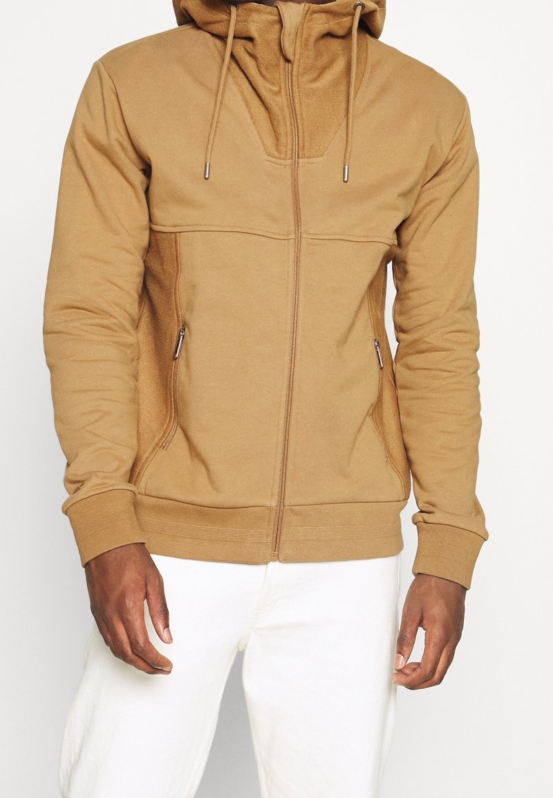 Homme portant un sweat à capuche zippé beige avec poches latérales sur un pantalon blanc, debout devant un fond uni.