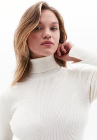 Weißer, gerippter Rollkragenpullover mit taillierter Passform, diagonal genähten Details und langen Ärmeln. Weiche Textur und leichtes Material.