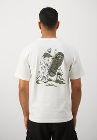 Hvid bomulds T-shirt med et grafisk motiv af en vandrer og et støvlemærke i grønt. Designet inkluderer bjerge i baggrunden.