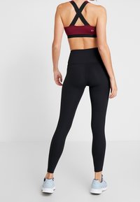 Burgundowy stanik sportowy z krzyżowym plecami w połączeniu z wysokim czarnym legginsami. Model nosi jasnoniebieskie buty sportowe. Gładka faktura tkaniny.