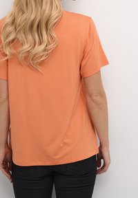 Triko s krátkým rukávem a kulatým výstřihem ve světle oranžové barvě, vyrobené z měkké látky. Má volný střih a rovný lem, nošeno s tmavými kalhotami.