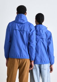 Chaquetas ligeras azules con capuchas, que cuentan con puños elásticos y un detalle en el yugo trasero, combinadas con pantalones de pana color tostado y pantalones de mezclilla claros.
