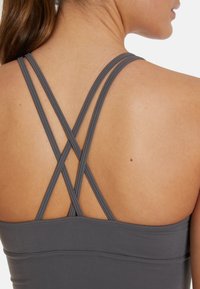 Siv tank top za šport z prepletenimi naramnicami na hrbtu, ki ima gladko teksturo in prilagojen dizajn. Poudarja ramena.