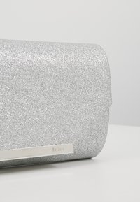 Stříbrná glitrová kabelka s hladkým povrchem a obdélníkovým tvarem. Obsahuje kovovou destičku s logem a design se sklápěcím uzavíráním.