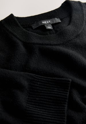 Črni pulover z okroglim izrezom, rebrastimi robovi na manšetah in spodnjem robu, narejen iz mehkega materiala. Vidna etiketa z napisom "NEXT", ki označuje znamko.