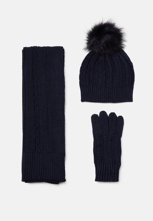 Ensemble d'hiver en maille bleu marine avec des motifs torsadés, comprenant un bonnet à pompon, des gants et une longue écharpe sur fond blanc.