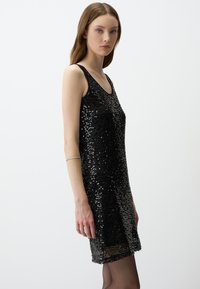 Robe noire sans manches recouverte de sequins, présentant un design ajusté et un décolleté arrondi, avec une bordure transparente à l'ourlet.