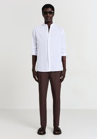 Camicia bianca con colletto alla cinese, abbinata a pantaloni aderenti marroni e sandali intrecciati neri. Tessuto morbido con una leggera texture.