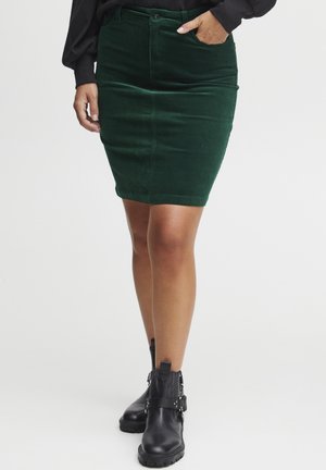 Mujer con falda a la rodilla de pana verde oscuro, botas negras hasta el tobillo y una camiseta negra de manga larga, con una mano en el bolsillo.