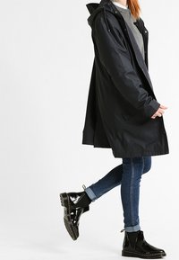 Czarna wodoodporna płaszcz przeciwdeszczowy z kapturem, bocznymi kieszeniami i luźnym krojem, zestawiony z niebieskimi dżinsami i błyszczącymi czarnymi botkami.