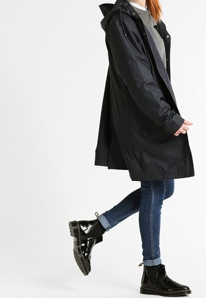 Czarna wodoodporna płaszcz przeciwdeszczowy z kapturem, bocznymi kieszeniami i luźnym krojem, zestawiony z niebieskimi dżinsami i błyszczącymi czarnymi botkami.