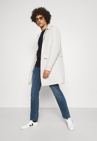 Light beige, lightweight coat with a collar, two front pockets, paired with dark jeans and white sneakers with black accents.