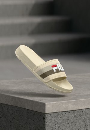 Μπεζ παντόφλα FILA με επενδυμένο λουράκι και λογότυπο, τοποθετημένη πάνω σε επιφάνεια από σκυρόδεμα με θολά σκαλοπάτια στο παρασκήνιο.