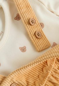 Gelber Cord-Romper mit Bärenmotiven, ausgestattet mit einer weißen Innenlage und braunen Knöpfen. Strukturierter Stoff mit gewellten Akzenten.