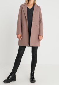 Lyserød, uldblandet oversized frakke med spidse revers og forreste lomme, parret med en grå sweater, sorte skinny jeans og sorte støvler.