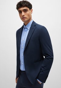 Navy blazer with a smooth texture, single-breasted design, and two front pockets, layered over a light blue button-up shirt.