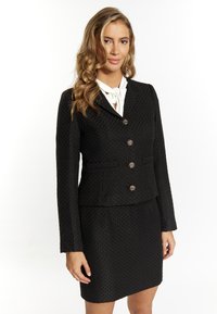 Blazer nero con texture, dal design aderente, caratterizzato da tre bottoni ornamentali, abbinato a una gonna aderente coordinata. Camicia bianca sotto.