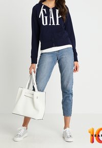 Navy zip-up hoodie met "GAP" logo, witte accenten, lichtblauwe cropped jeans, witte tote bag en zilveren sneakers.