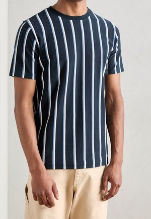 Mężczyzna w granatowym krótkim T-shircie z pionowymi białymi paskami i beżowych spodniach, stojący na jednolitym tle.
