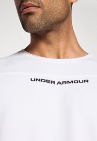 Άσπρο βαμβακερό μπλουζάκι με στρογγυλή λαιμόκοψη, με μαύρο λογότυπο "UNDER ARMOUR" στο στήθος, λεία υφή και casual σχεδίαση.