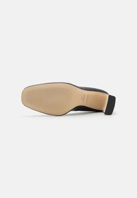 Chaussure à talons noire avec une tige en cuir lisse et une semelle en caoutchouc texturé. Présente un talon carré et un logo de marque en relief sur la semelle intérieure.