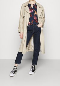 Trench coat beige, top navy con fiori e scollatura, jeans in denim scuro e sneakers nere con suola e lacci bianchi.