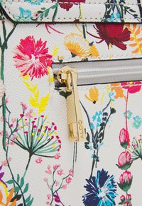 Sac à motif floral avec une surface blanche texturée. Comprend une tirette de fermeture éclair dorée avec "ALDO" gravé. Fleurs colorées en rose, jaune et bleu.