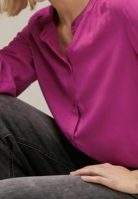 Blusa fucsia con bottoni sul davanti, maniche lunghe e collo alto; tessuto liscio abbinato a jeans grigio scuro.