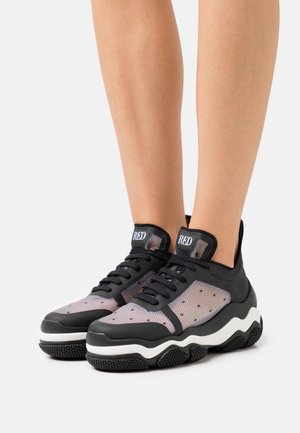 Pieds chaussés de baskets en maille noire et grise avec des semelles épaisses blanches et noires texturées et une étiquette "RED" sur la languette, présentés sur un fond blanc.
