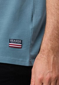 Męska koszulka z krótkim rękawem w miękkim niebieskim kolorze, z granatową i czerwoną naszywką na dolnym brzegu z napisem „SEASIDE NAUTICAL LEGACY.”