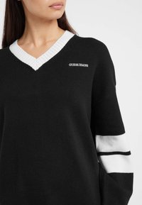 Pull en tricot noir avec un col en V côtelé blanc, présentant un logo sur la poitrine et des rayures blanches sur les manches. Texture douce, coupe décontractée.