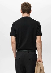Tricou negru cu mânecă scurtă, gât clasic, textură netedă și design ajustat, purtat cu pantaloni negri pe formă și ținând o jachetă bej.