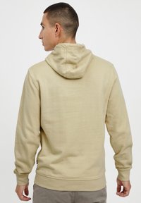 Beige mikina s klokaní kapsou, kapucí na šňůrku a žebrovanými manžetami a lemem, vyrobená z měkké, strukturované tkaniny. Zobrazeno zezadu.