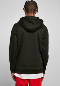 Sweatshirt à capuche noire avec une texture douce, des poignets et un ourlet côtelés. Porté par-dessus un look superposé blanc, présenté de dos.