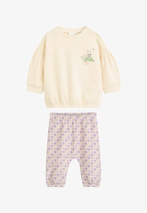 Lys beige sweatshirt med pufærmer og et cartoon-grafik, parret med lavendelternede bukser med et blomstermotiv.