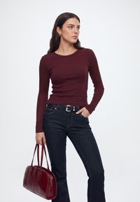 Femme aux longs cheveux foncés portant un haut bordeaux à manches longues et un jean sombre, tenant un sac à main bordeaux assorti dans une main.