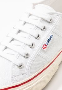 Białe płócienne trampki z okrągłymi, srebrnymi oczkami, białymi sznurowadłami, naszywką logo w czerwono-niebieskich kolorach oraz teksturowaną gumową podeszwą z czerwoną linią.