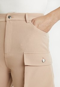 Beige kalhoty s postranní kapsou a přední kapsou na ruce, s kovovým knoflíkem a rukou částečně zasunutou v kapse.