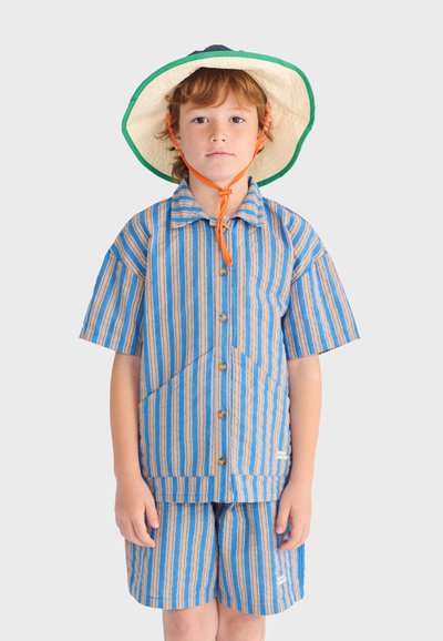 Enfant portant une chemise à manches courtes rayée bleu et beige et un short assorti, avec un chapeau à large bord maintenu par une sangle orange sous le menton.