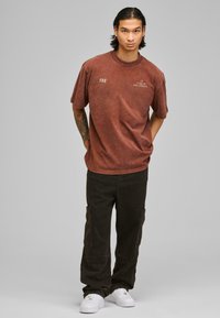 Camiseta oversized marrón con una textura desgastada y texto impreso, combinada con pantalones cargo oscuros y zapatillas blancas. Ropa simple y casual.