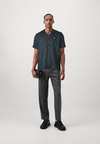 Camisa de cuadros negra de manga corta con líneas azules, con cierre de botón y un logo amarillo, combinada con pantalones gris ajustados y zapatos negros.