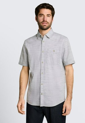 Mann mit kurzen dunklen Haaren, der ein hellgraues kurzärmeliges Hemd mit Knöpfen und dunkle Hosen trägt, steht vor einem einfarbigen Hintergrund.