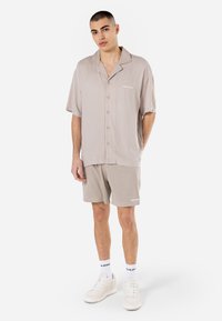 Kortærmet beige skjorte med knaplukning og notch-krave, kombineret med lette mesh-shorts. Hvide sneakers og strømper med logo-detaljer fuldender looket.