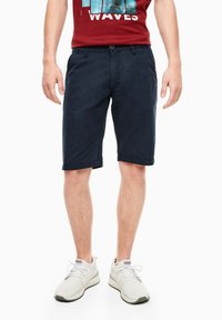 Muž v námořnicky modrých šortkách po kolena, bílých teniskách a červeném tričku s potiskem, stojí s uvolněnýma rukama podél těla.