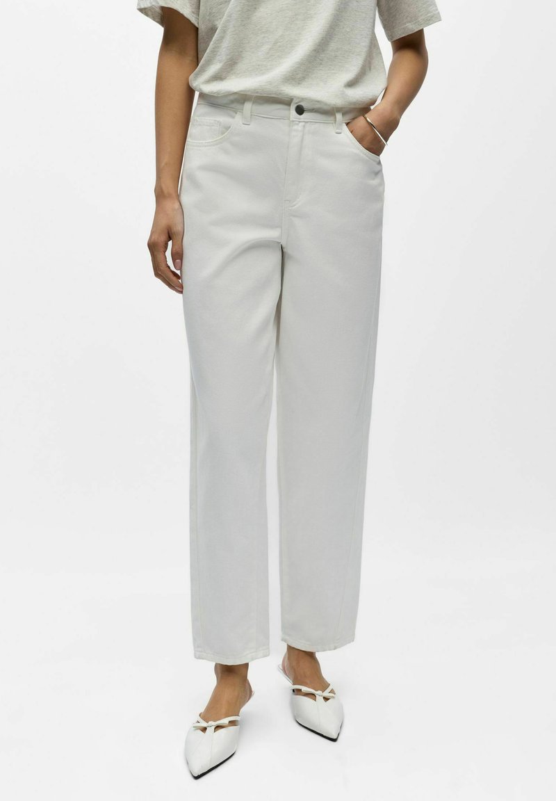 Personne portant un pantalon blanc taille haute, jambe droite, un haut gris clair à manches courtes, des ballerines blanches pointues avec des accents de nœud, et un bracelet argenté.