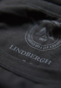 Zwarte stof met een gladde structuur, met daarop een logo en de naam "LINDBERGH" in lichtgrijs gedrukt. Afgeronde randen in het ontwerp.