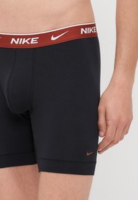 Sorte bomulds boxer shorts med en bordeaux elastisk talje med hvid "NIKE" branding og et lille rødt logo på siden af benet.