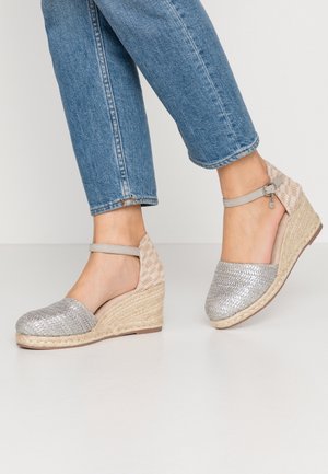 Person iført blå jeans og sølvfarvede espadrille-kilehøjhælede sko med ankelstropper, stående på en lys gulv mod en ensfarvet baggrund.