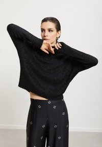 Černý, krátký svetr s volným střihem, texturovanými metalickými vlákny a širokými rukávy. V kombinaci s černými kalhotami s geometrickým vzorem.